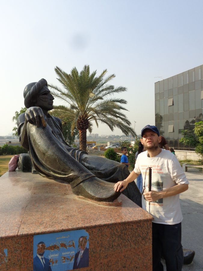 Tony touching a bronze statue of King Shahryar wearing traditional Middle Eastern clothing and a turban. He is sculpted reclining on a reddish marble base. This is located alongside the Queen Scheherazade sculpture shown in the previous photo. In the background a palm tree can be seen and there is a modern glass-clad building to the right. The far bank of the river Tigris can be seen away in the distance.