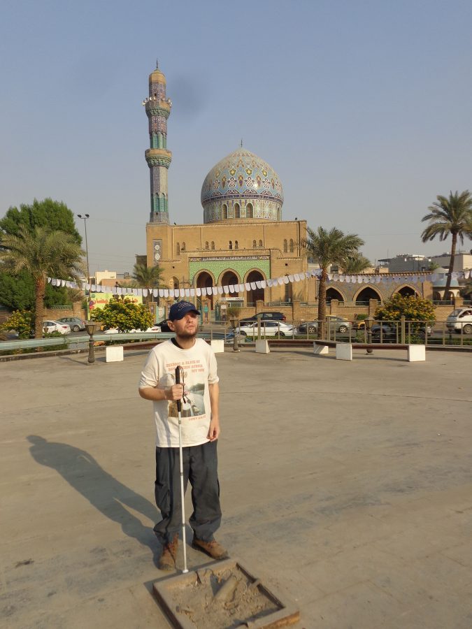 Tony in Al-Firdos Square with the 17th of Ramadan Mosque in the background. The mosque has a large, ornate blue, turquoise and yellow dome with intricate geometric patterns and a tall minaret decorated with similar colourful tilework. The building was completed in 1959. Al-Firdos Square is a public square close to the Ishtar and Palestine Hotels in central Baghdad. It is named after the Persian word Ferdows meaning 'paradise'. This square is where the large statue of Saddam Hussein, then President of Iraq, was toppled by a US Marine military vehicle on 9th April 2003.