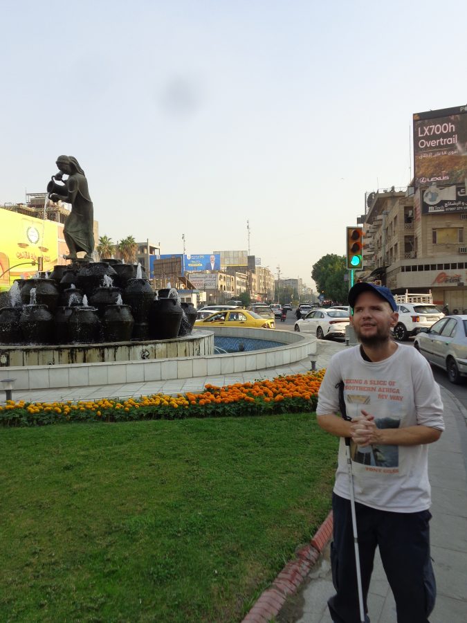 Tony again stood next to the Kahramana fountain. There are orange and yellow flowers planted in front.  The fountain is in the middle of a junction between busy roads. Cars can be seen queuing around the side. There are several large billboards in view attached to surrounding buildings.