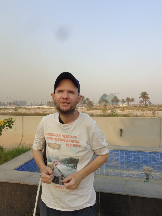 Another view of the Al-Shaheed Monument with Tony in the foreground. The monument includes a 40-metre tall turquoise dome with a pointed top. The dome is split down the middle into two off-set parts with an eternal flame in the middle. The monument is on a circular platform 190 metres in diameter in the centre of an artificial lake: the lake is not visible in the photo. A museum, library, cafeteria, lecture hall, and exhibition gallery are on two levels underneath the domes. The dome is in the middle distance with scattered palm trees towards the foreground.
