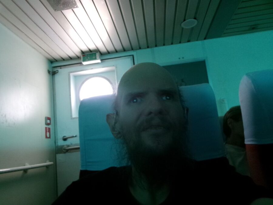 Tony onboard the Limni Express ferry travelling from the Greek island of Lemnos (Limnos) to Canakkale, Türkiye. He is sitting at the end of a row of high-backed seats with white headrest covers. There is an emergency exit doorway behind to the left.