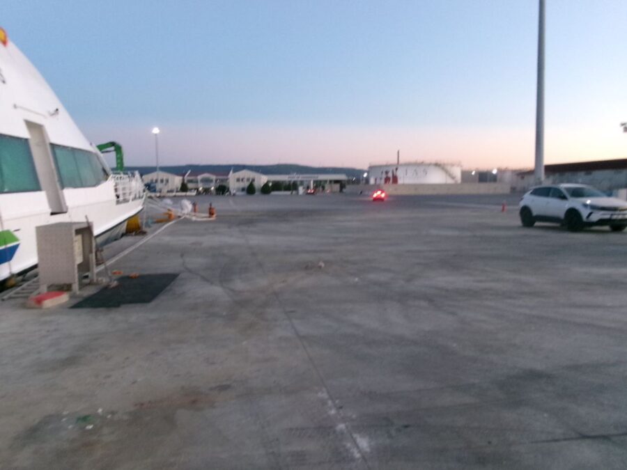 At the ferry port at Kepez, Canakkale, Türkiye. The photo is taken at dusk. A large open concrete dock area extends in front with the small white ferry docked to the left. Various buildings and a large white storage tank can be seen around the port area.