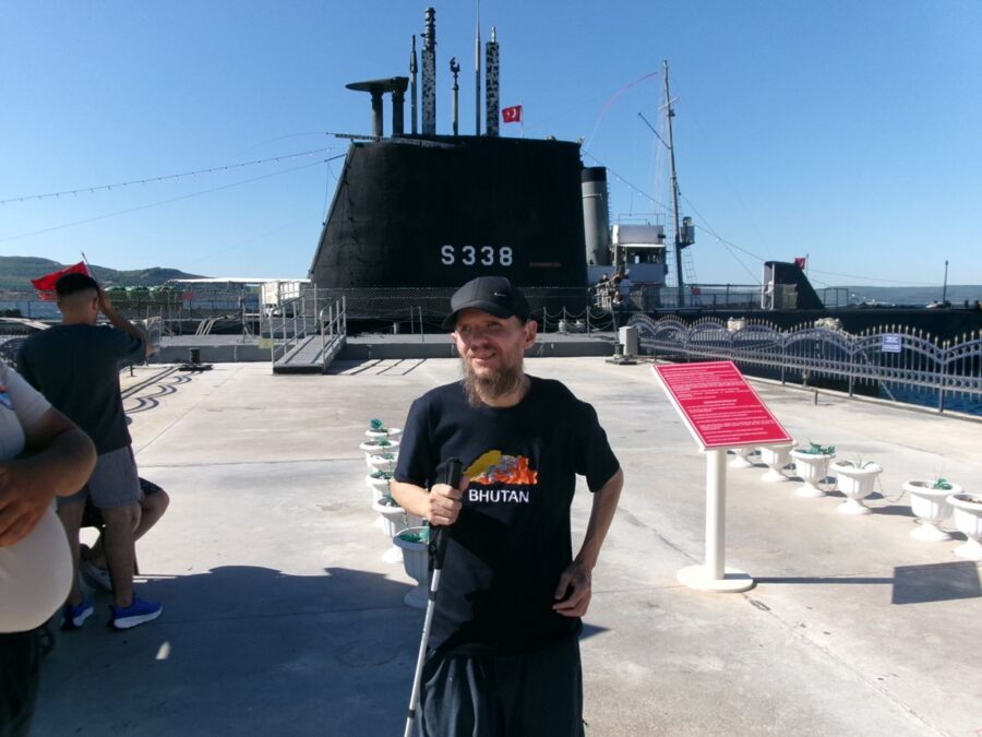 A smiling Tony stood on an outdoor concrete deck in front of a large black submarine marked “S338”. Tony is wearing a black baseball cap, a navy blue T‑shirt with the Bhutanese flag and the word “Bhutan” printed on it. He is holding his white cane in his right hand and his left hand is resting on his hip. The submarine has several vertical masts and periscopes rising from its central tower as well as a small Turkish flag. The submarine was originally known as USS Thornback and was built and launched in 1944. It was struck from the US Navy's register in 1973, and was then transferred to the Turkish Naval Forces, and renamed TCG Uluçalireis (S 338), serving until 2000. She has since become a museum ship at the Çanakkale Military Marine Museum.