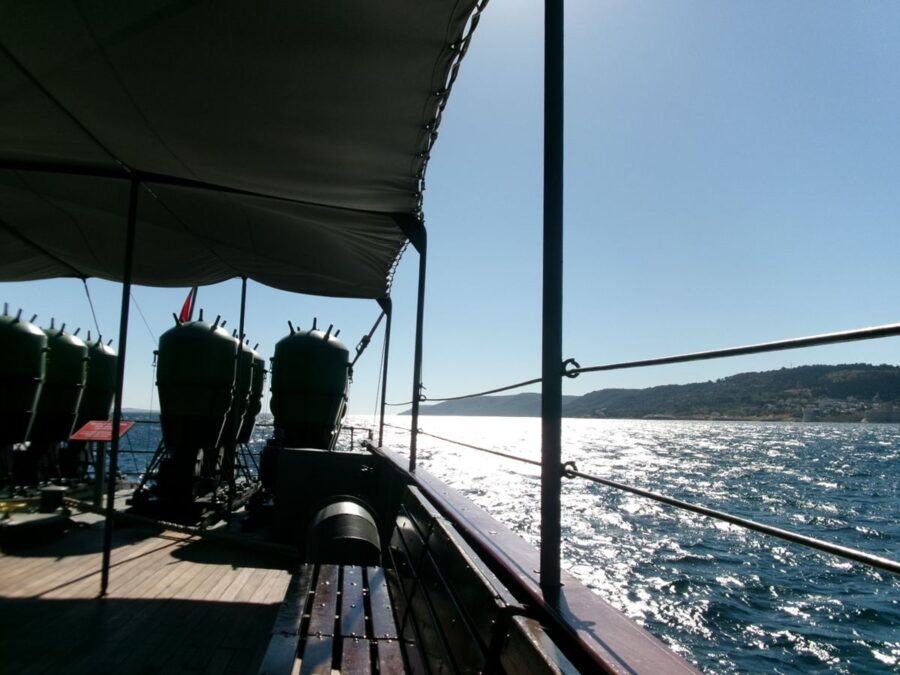 Onboard a replica of the Ottoman Navy minelayer ship Nusret at Çanakkale Military Marine Museum (Naval Museum). The original ship was commissioned in 1913 and laid mines that sank three British warships and badly damaged three others in March 1915 during World War I. The ship then served various roles in the Turkish Navy before being sold to commercial interests in 1962. In April 1989, she capsized and sank near Mersin Harbour, and lay there for 10 years before being salvaged in 1999. Nusret was then converted into a museum ship in Mersin, and additionally this replica was built, and opened as a museum ship in Çanakkale in 2011. The photo is taken on the deck. In front are rows of large barrel shaped objects, probably replica sea mines. The Dardanelles extends out ahead lit by the sun reflecting on the sea.