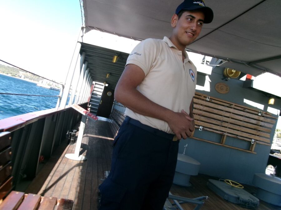 Again on the deck of the replica mine-laying ship, Nusret, moored at Çanakkale Military Marine Museum on the Dardanelles strait. From the deck, the deep blue sea can be seen, lit by the sun. In front a man is stood dressed in a baseball cap and a beige polo shirt. He is one of the museum's staff members and guides. The deck is shaded by a canvas canopy. The floor of the deck is finished with wooden planks and a wooden bench is visible to the left.