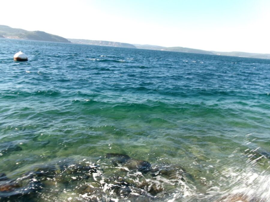 Clear blue-green water at the rocky shoreline in Çanakkale city centre. Splashes from small waves can be seen. A white and red buoy is floating in the water ahead. In the distance cliffs and hills on the opposite side of the Dardanelles can be seen. At its narrowest point the strait is about 1 mile (1.6km) wide. The sky is slightly hazy giving it a white to pale blue colour.