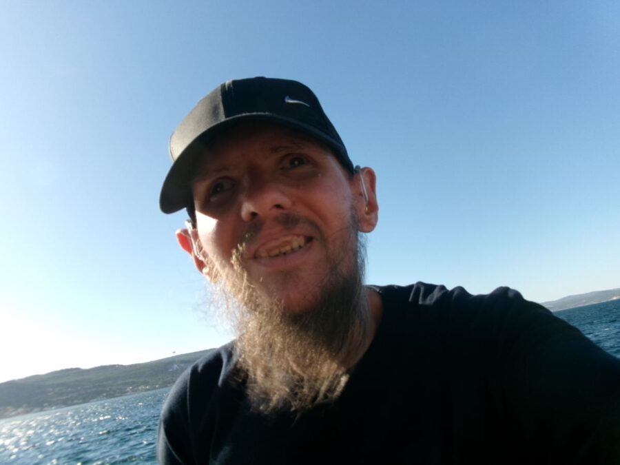 Tony smiling in the foreground. He is wearing a dark cap with the sea behind. Land on the opposite side of the Dardanelles is visible in the distance. Photo taken close to the Military Marine Museum in Çanakkale.