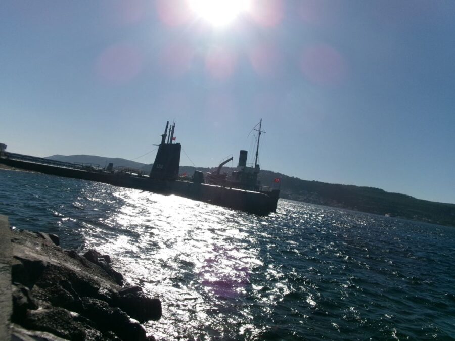 Looking out across the Dardanelles from Çanakkale. The historic submarine TCG Uluçalireis (S338) can be seen ahead. The submarine and the water across the strait are illuminated by the sun. The Dardanelles is a narrow, natural strait, which forms part of the continental boundary between Europe and Asia. It is also an internationally significant waterway joining the Aegean and Mediterranean seas with the Sea of Marmara with onward access to the Black Sea.