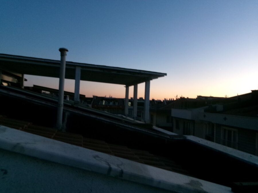 View across rooftops at dusk. The sky is a clear blue. Ahead the sun is almost set with the horizon lit up in a gradient of red-orange through to yellow. Taken from the balcony of Tony's Turkish friend's house in Çanakkale. The surrounding roofs are silhouettes with chimneys, satellite dishes, and other structures visible.