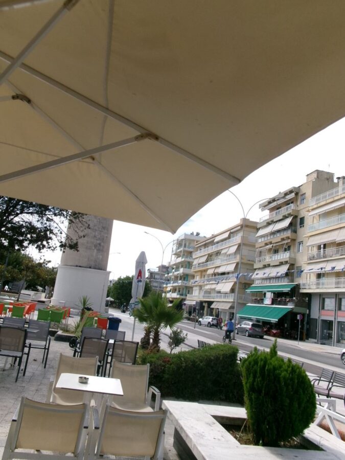 Outdoor café terrace with a large cream-coloured awning overhead. Located on the sea front promenade in Alexandroupolis. The base of Alexandroupolis Lighthouse can be seen directly ahead. The promenade is lined with trees and a road runs along side. It appears quiet with few people in view.